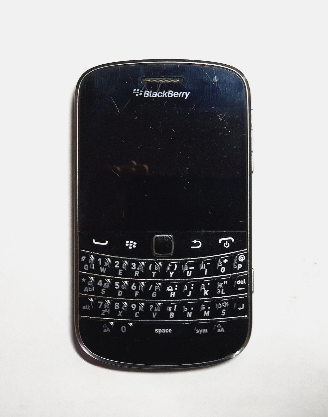 Front view of a black BlackBerry Bold 9900 smartphone with its screen off and a physical QWERTY keyboard marked in Latin and Cyrillic letters
